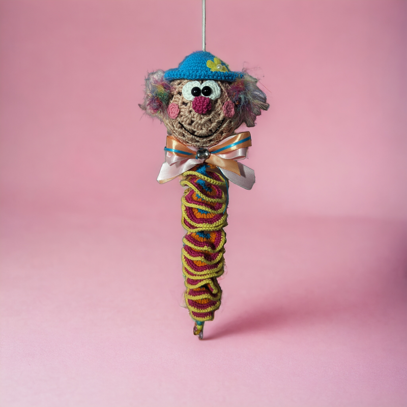 gehäkelter Clown‑Anhänger mit blauem Häubchen und buntem Spiralkörper vor rosa Hintergrund