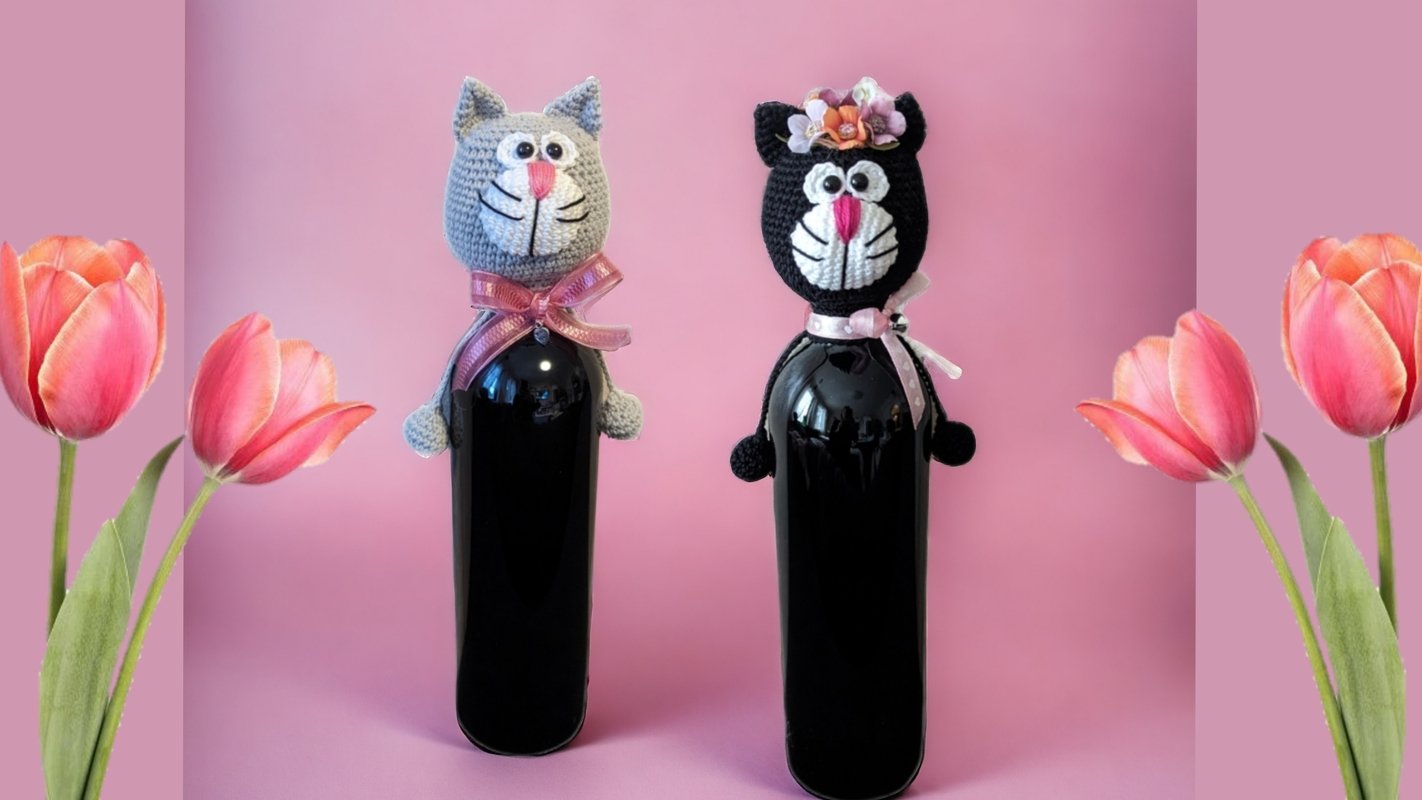 Zwei Flaschen mit gehäkelten Katzenköpfen vor rosa Hintergrund, links grau mit rosa Schleife, rechts schwarz mit Blumenkrone.