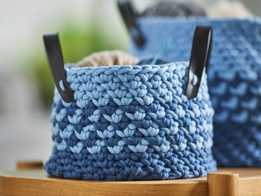 Nahaufnahme eines runden, gehäkelten Korbs in Hell- und Dunkelblau mit zwei dunklen Griffen, gefüllt mit hellem Garn und einer Häkelnadel auf einem Holztisch.