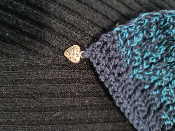 Nahaufnahme Ecke eines gehäkelten Dreieckstuchs in Blau-Türkis mit kleinem Metallherz auf schwarzem Rippstoff