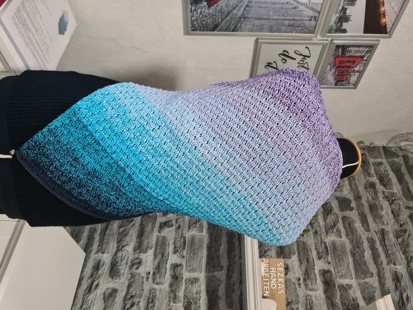 Gehäkeltes Dreieckstuch mit Farbverlauf von Petrol über Türkis zu Lila auf einer Schaufensterpuppe vor einer Ziegelwand.