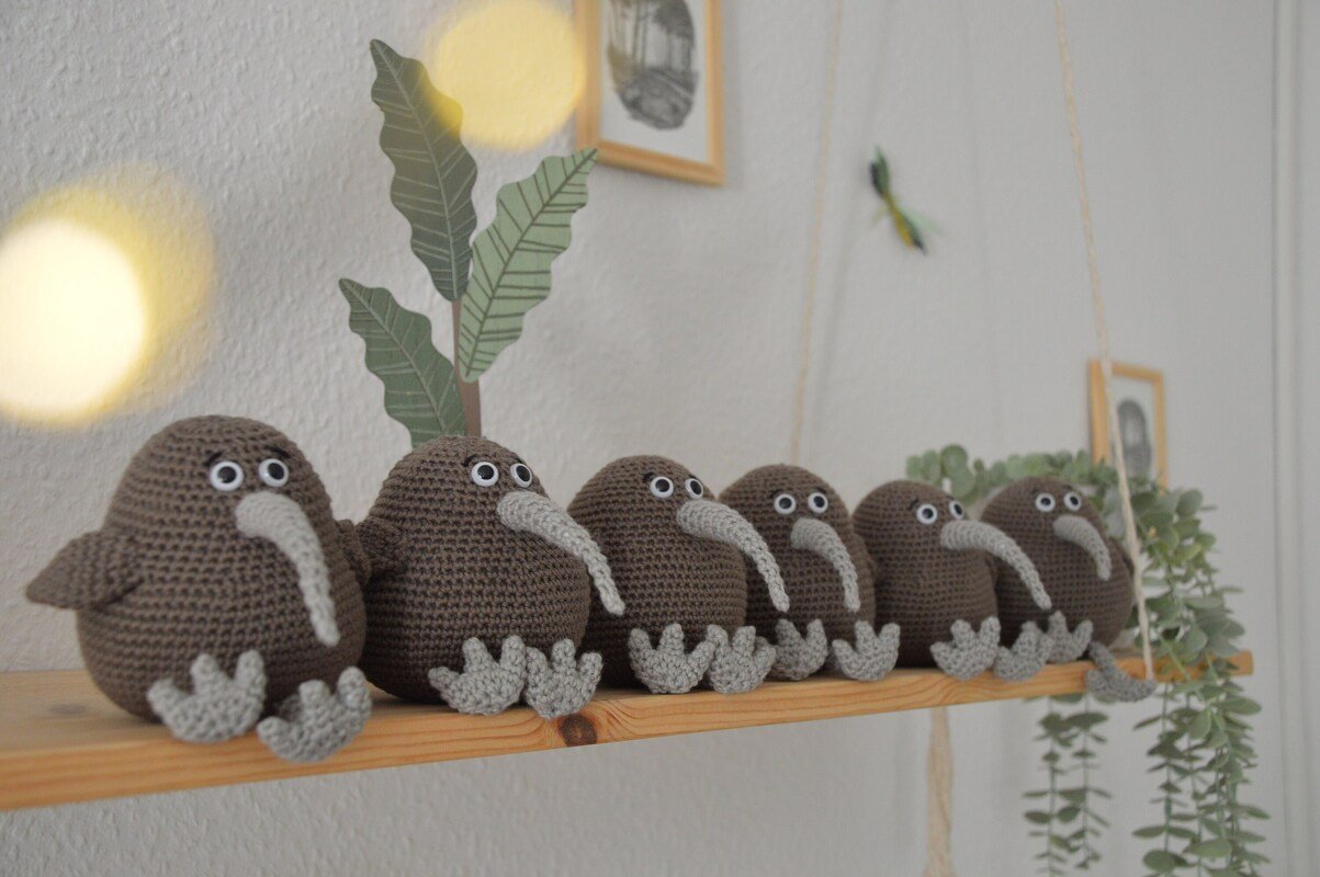 Sechs gehäkelte braune Amigurumi-Kiwis mit hellgrauen Schnäbeln und Füßen auf einem Holzregal.