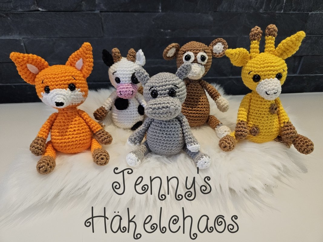 Fünf gehäkelte Amigurumi-Tiere (Fuchs, Kuh, Nilpferd, Affe, Giraffe) sitzend auf weißem Fell vor dunklem Hintergrund