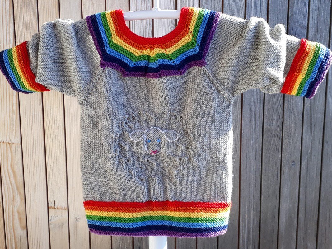 Light grey knitted baby sweater with rainbow bands on collar, cuffs, and hem and a textured sheep motif on the front.