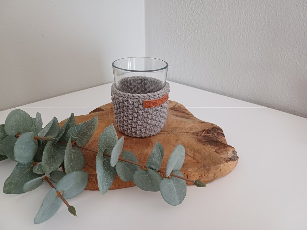 Klares Glas mit grauem gehäkeltem Überzug auf Holzbrett neben Eukalyptuszweig