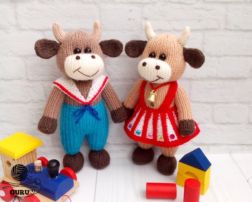 Two handmade knitted amigurumi cows: one wearing blue sailor overalls, the other in a red beaded dress with a small bell, posed with wooden blocks on a white surface.