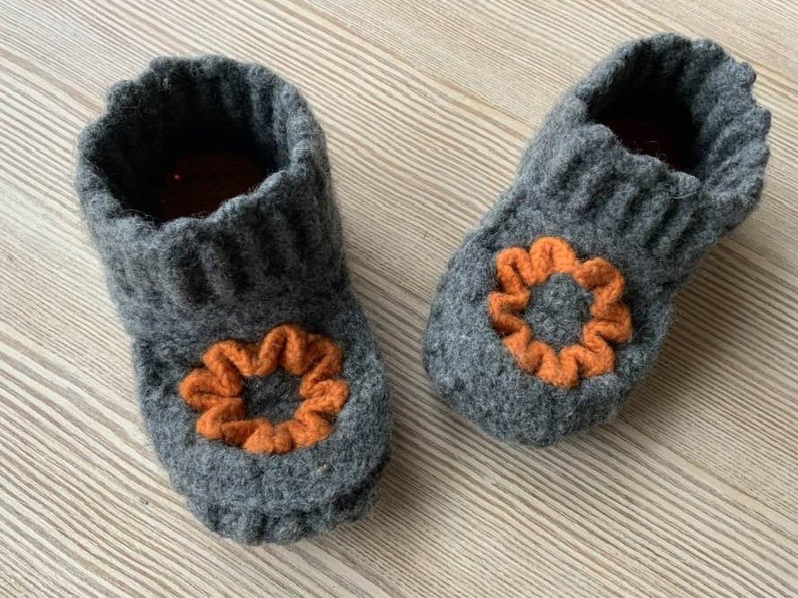 Zwei dunkelgraue gehäkelte Hausschuhe mit orangefarbenen Blütenapplikationen auf hellem Holzboden.