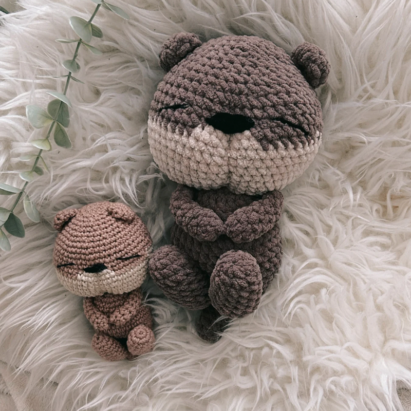 Two crocheted otter amigurumi in brown tones on a white faux-fur background