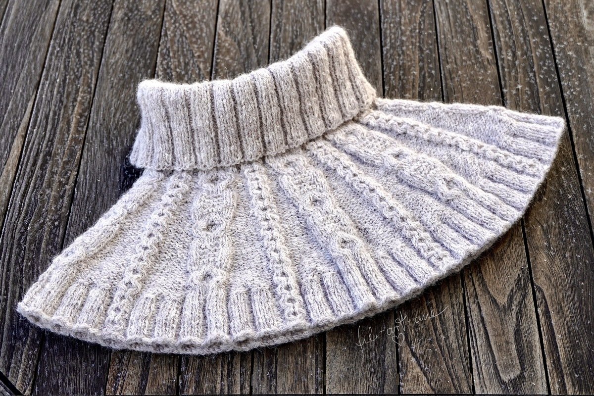 Close-up of a light beige knitted collar scarf with cable panels and a ribbed neck, laid on a dark wood surface.