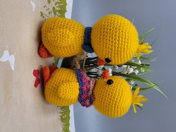 Zwei gelbe, gehäkelte Küken mit bunten Schals neben Maiglöckchen auf beigem Untergrund.