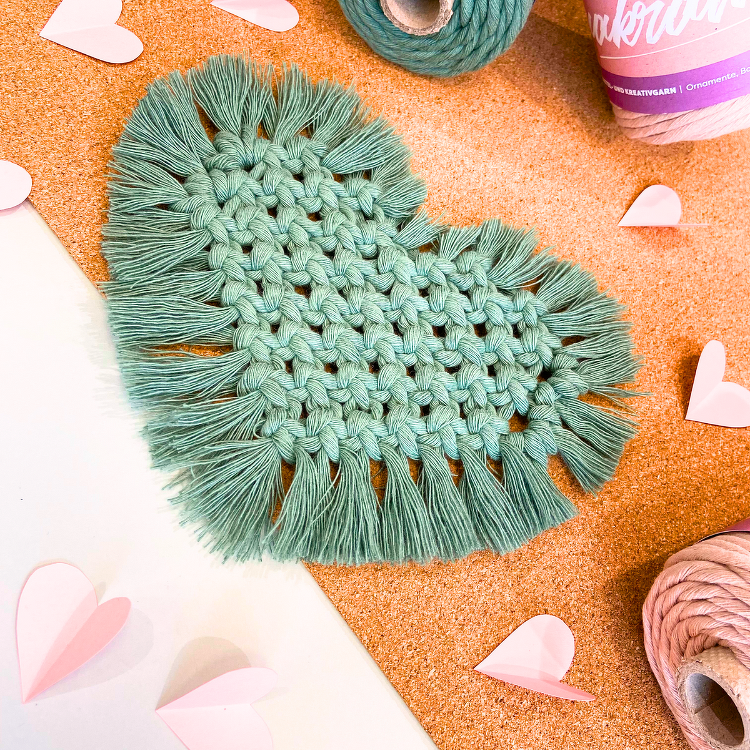 Nahaufnahme eines salbeigrünen Makramee-Herzens mit sichtbaren Knotenreihen und Fransen auf Korkuntergrund