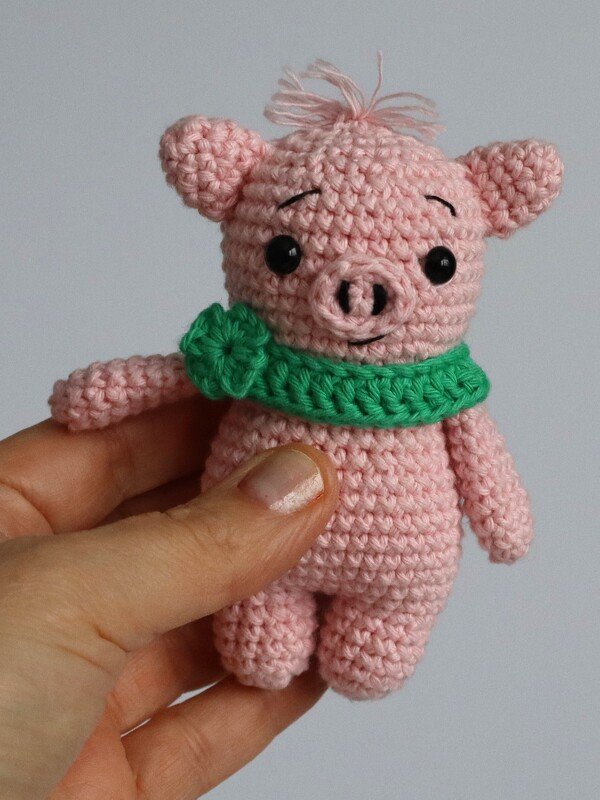 Rosafarbenes gehäkeltes Amigurumi-Schweinchen mit grünem Schal und Blüte, in einer Hand gehalten