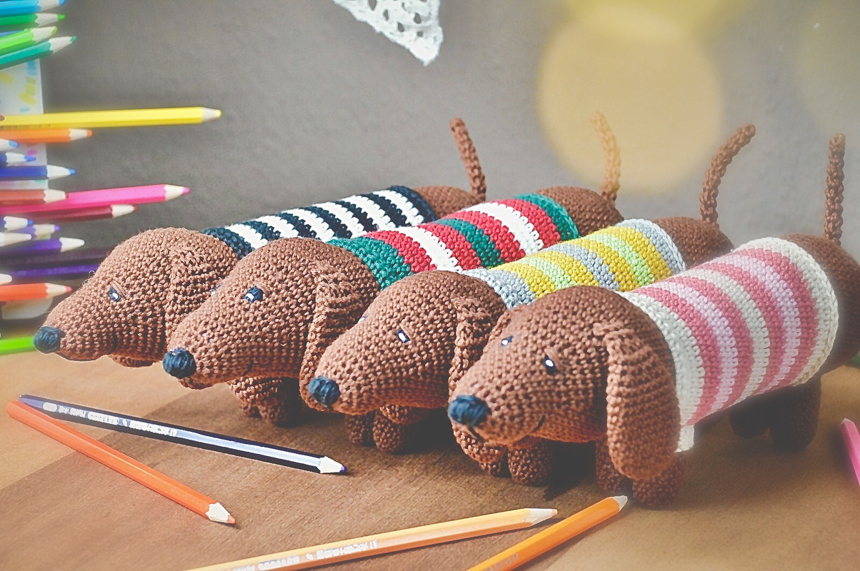 Vier gehäkelte Dackel (Amigurumi) mit gestreiften Pullovern auf Holztisch, umgeben von Buntstiften