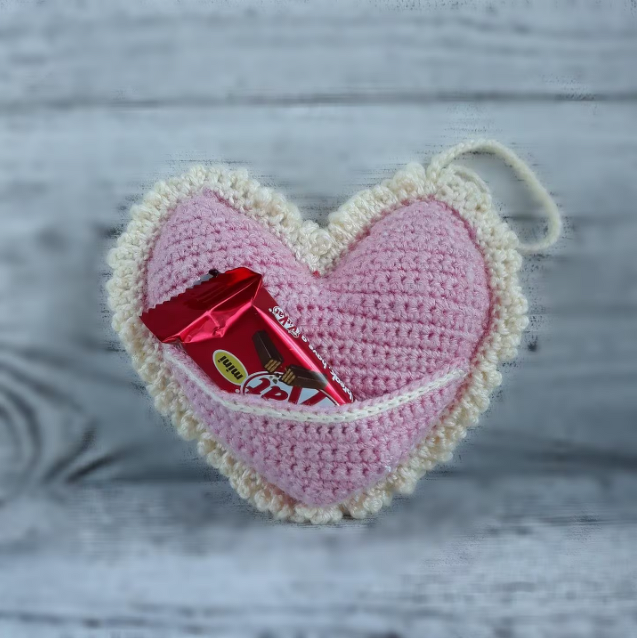 Pink crocheted heart with cream ruffled edge and front pocket holding a small wrapped treat.