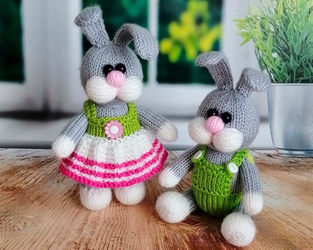Two grey hand-knitted amigurumi bunnies—one standing in a dress, one sitting in green overalls—on a wooden surface.