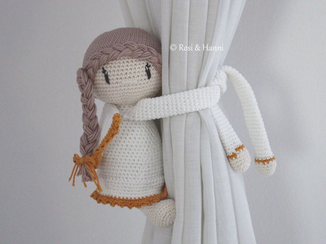 Gehäkeltes Amigurumi-Engelchen um einen weißen Vorhang geschlungen, mit geflochtener Zopf und senffarbenen Saumdetails