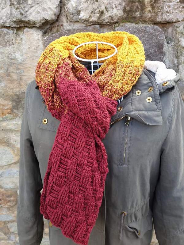 Gehäkelter Schal in Gelb, Orange und Rot mit strukturiertem Muster, drapiert an einer Schaufensterpuppe vor einer Steinwand.