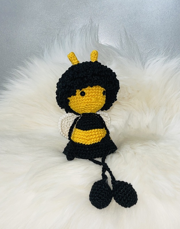 Gehäkelte schwarz-gelbe Amigurumi-Biene mit Lockenkranz und weißen Flügeln auf flauschigem Stoff.