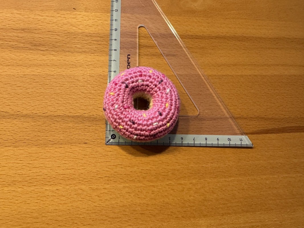 Kleiner rosa gehäkelter Donut neben transparentem Dreiecklineal auf Holz