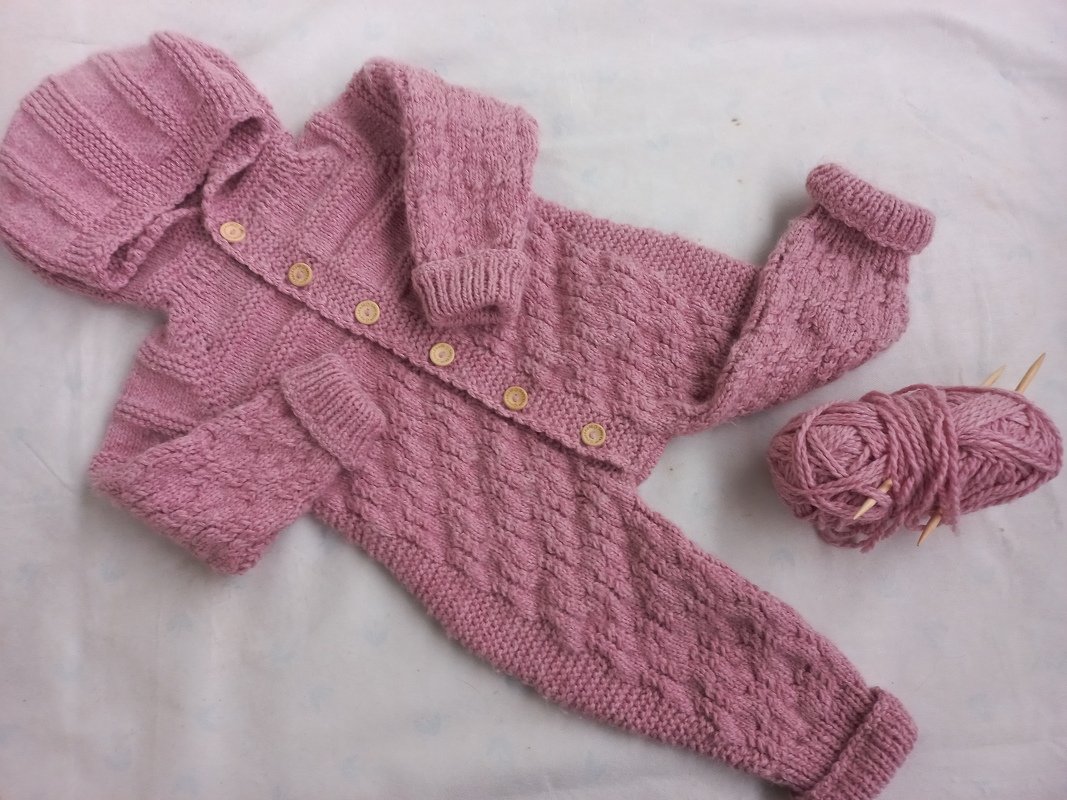 Altrosa gestrickter Baby-Overall mit Kapuze, Knöpfen und Zopfmuster, daneben Wollknäuel und Stricknadeln