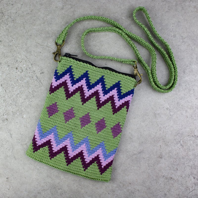 Grüne gehäkelte Umhängetasche mit Zickzack- und Rautenmuster in Lila und Blau auf grauem Untergrund
