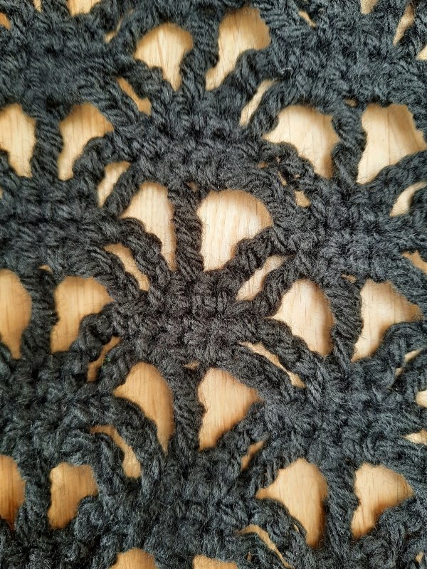Nahaufnahme eines dunkelgrauen gehäkelten Schals mit Lochmuster auf Holzuntergrund.