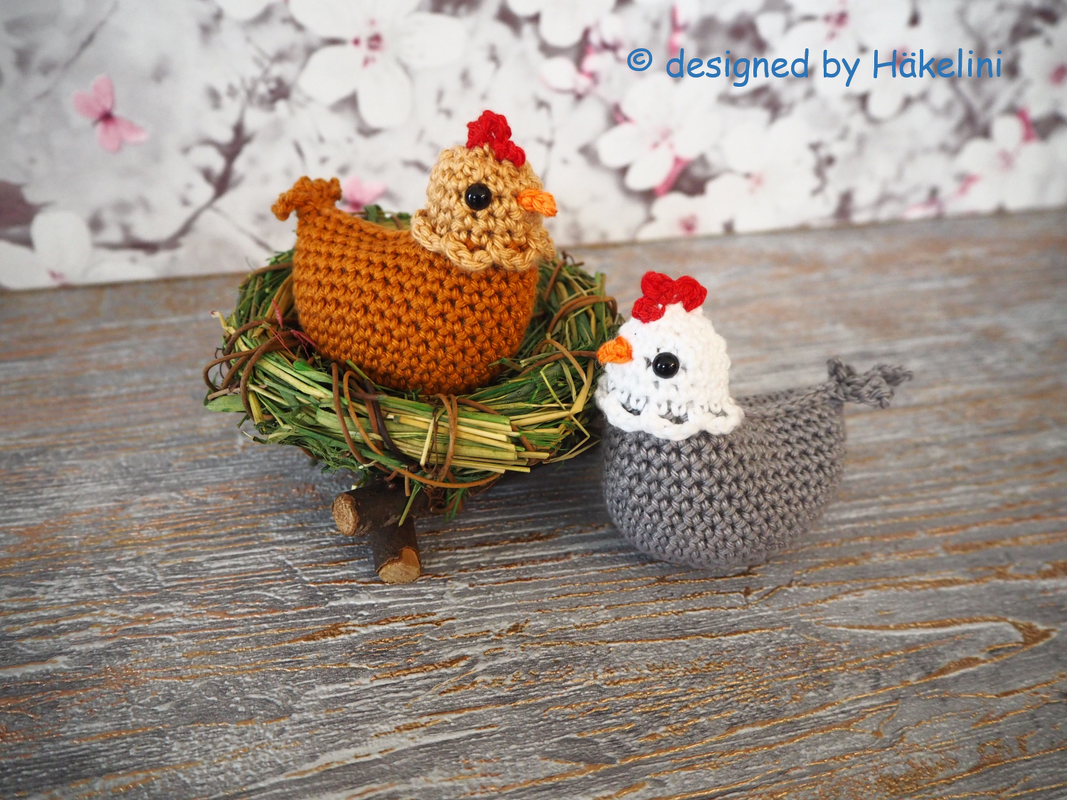 Zwei gehäkelte Amigurumi‑Hühner, eines braun im Strohnest, das andere grau auf einer Holzoberfläche
