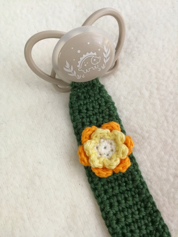 Dunkelgrüne gehäkelte Schnullerkette mit gelb-orangener Häkelblume an beigem Schnuller mit Igel-Motiv auf hellem Hintergrund.