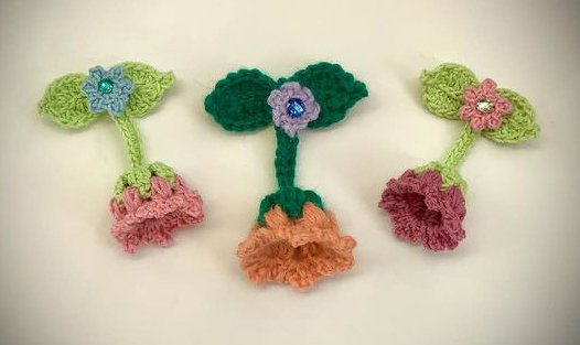 Drei gehäkelte Blumenapplikationen in Rosa, Orange und Magenta mit grünen Blättern und kleinen Glitzersteinen.