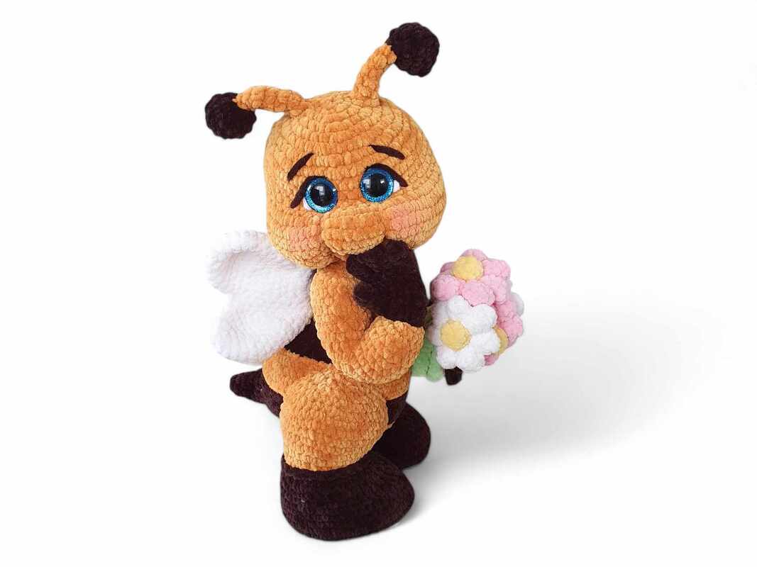 Orange and brown crocheted bumblebee amigurumi with blue eyes and white wings holding a small bouquet of pink and white yarn flowers.