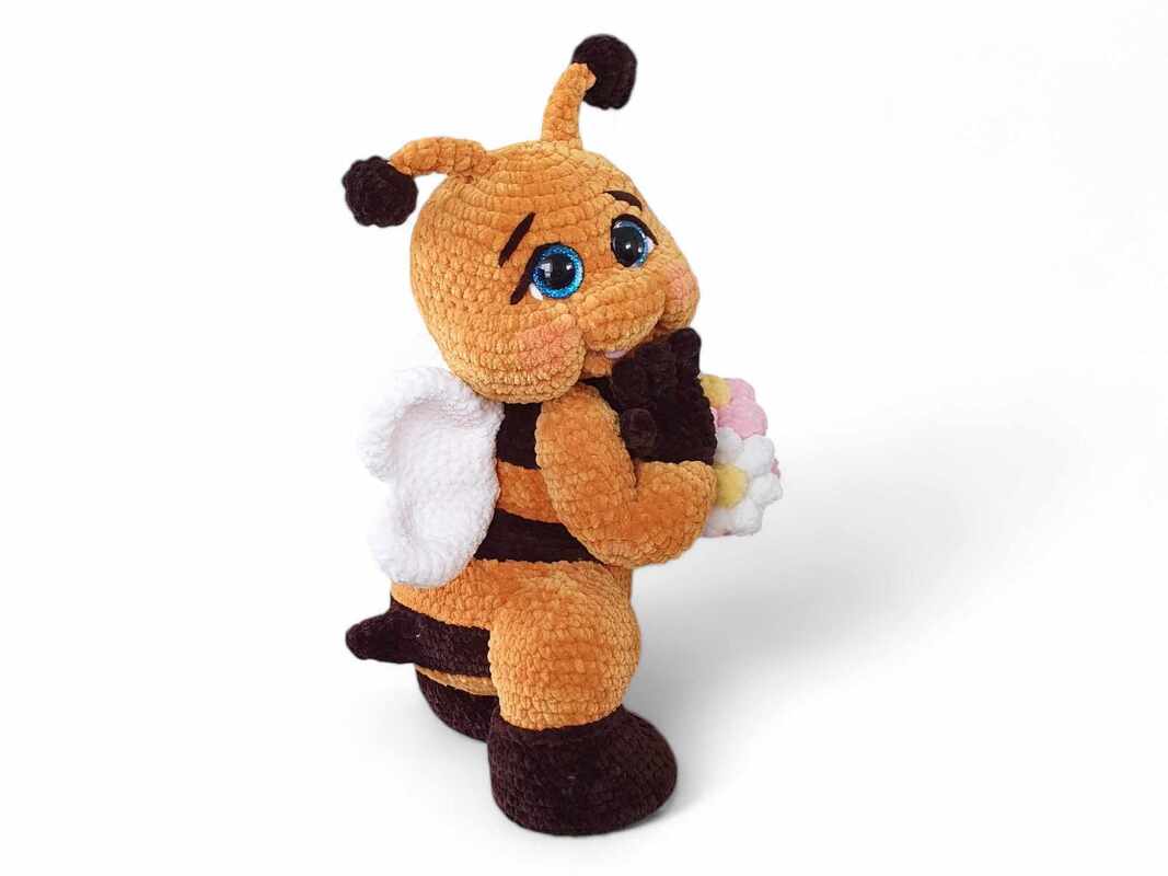 Amigurumi bee toy with orange and brown stripes, white wings, blue eyes, holding a small bouquet of white, pink, and yellow flowers.