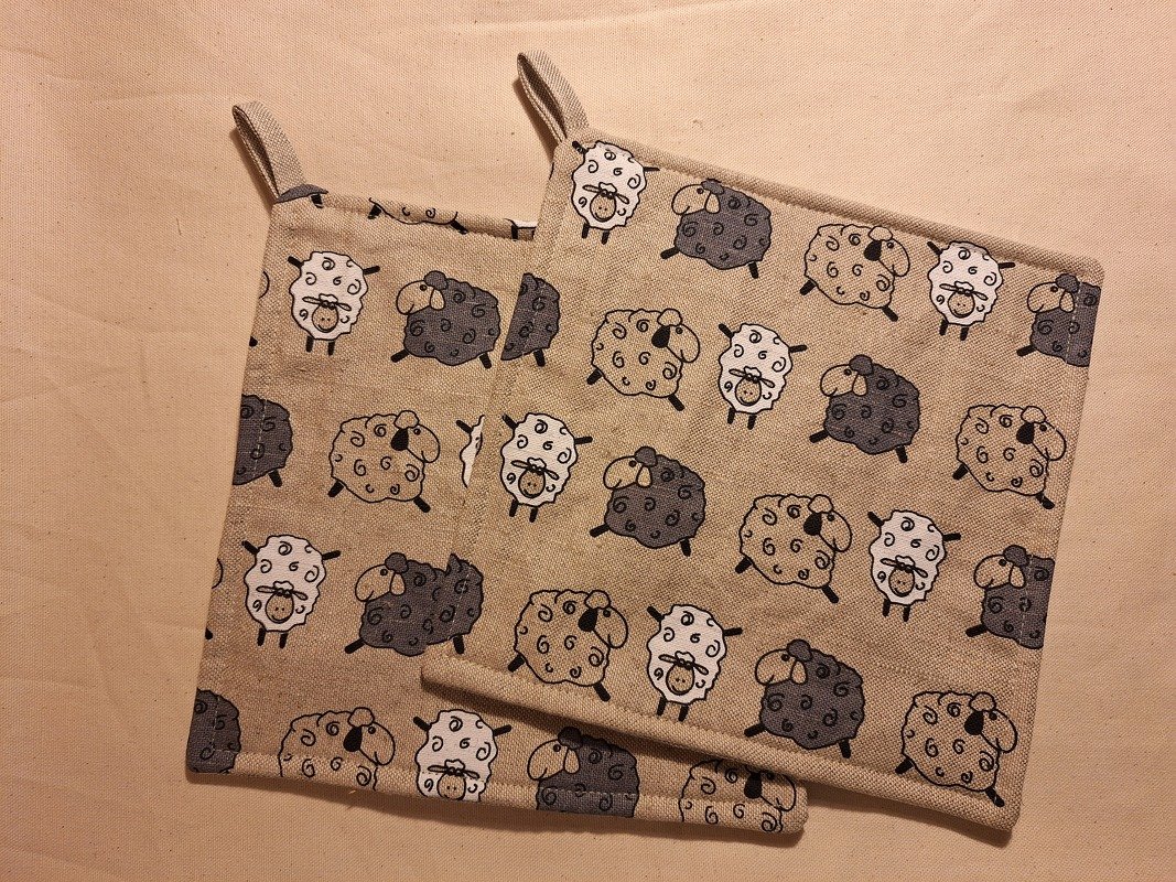 Zwei quadratische Topflappen aus beigem Stoff mit kleinem Schafsmuster und Aufhängeschlaufen
