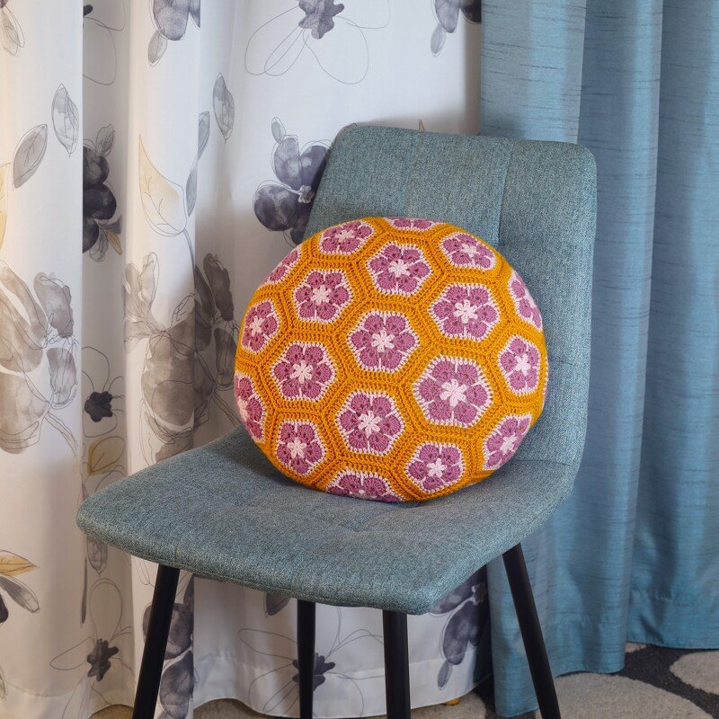 Rundes gehäkeltes Kissen mit African Flower-Motiven in Orange und Pinktönen auf einem Stuhl.