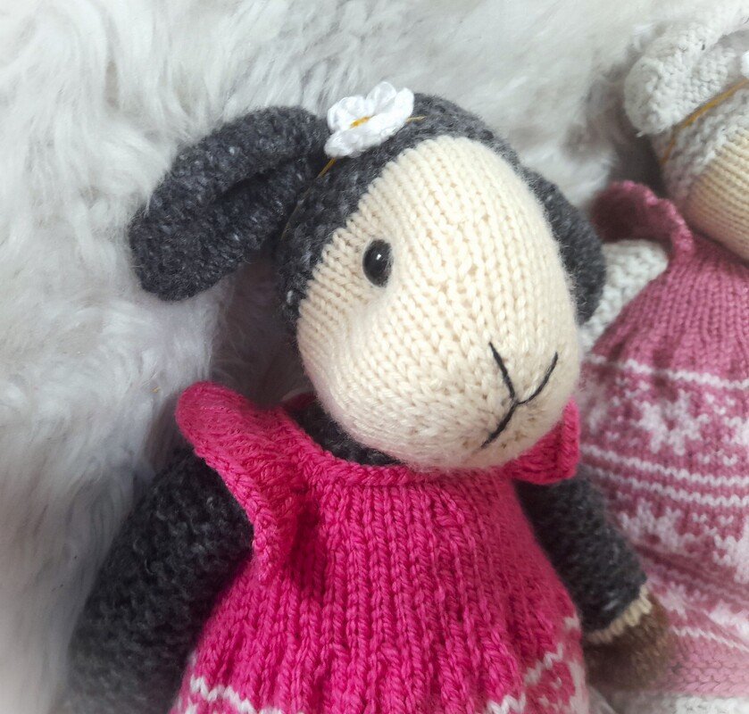 Gestricktes Amigurumi-Lamm mit rosa Kleid, grauen Ohren und Blüte im Haar auf hellem Hintergrund
