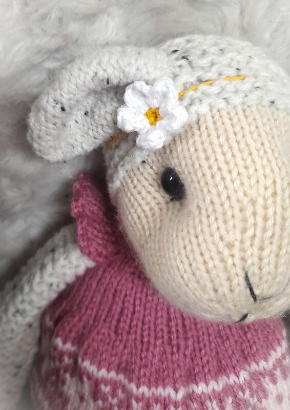 Knitted lamb amigurumi with a white crocheted flower on its head and a pink patterned sweater.
