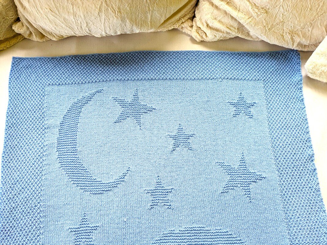 Pale blue rectangular knitted baby blanket showing a crescent moon and stars worked in textured stitches with a bordered edge.