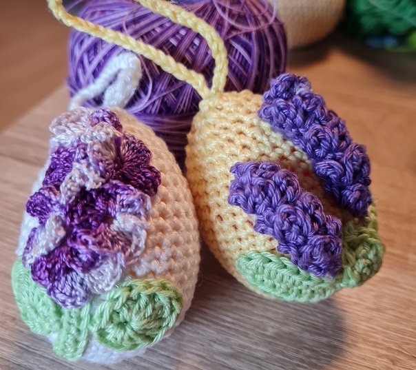 Zwei gehäkelte Ostereier in Pastellfarben mit lila Blumenapplikationen und grünen Details, mit Aufhängeschlaufe.