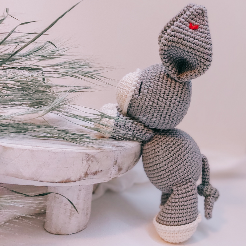 Gehäkelter grauer Amigurumi-Hase mit cremefarbenen Pfoten und rotem Herz auf dem Ohr, seitliche Ansicht