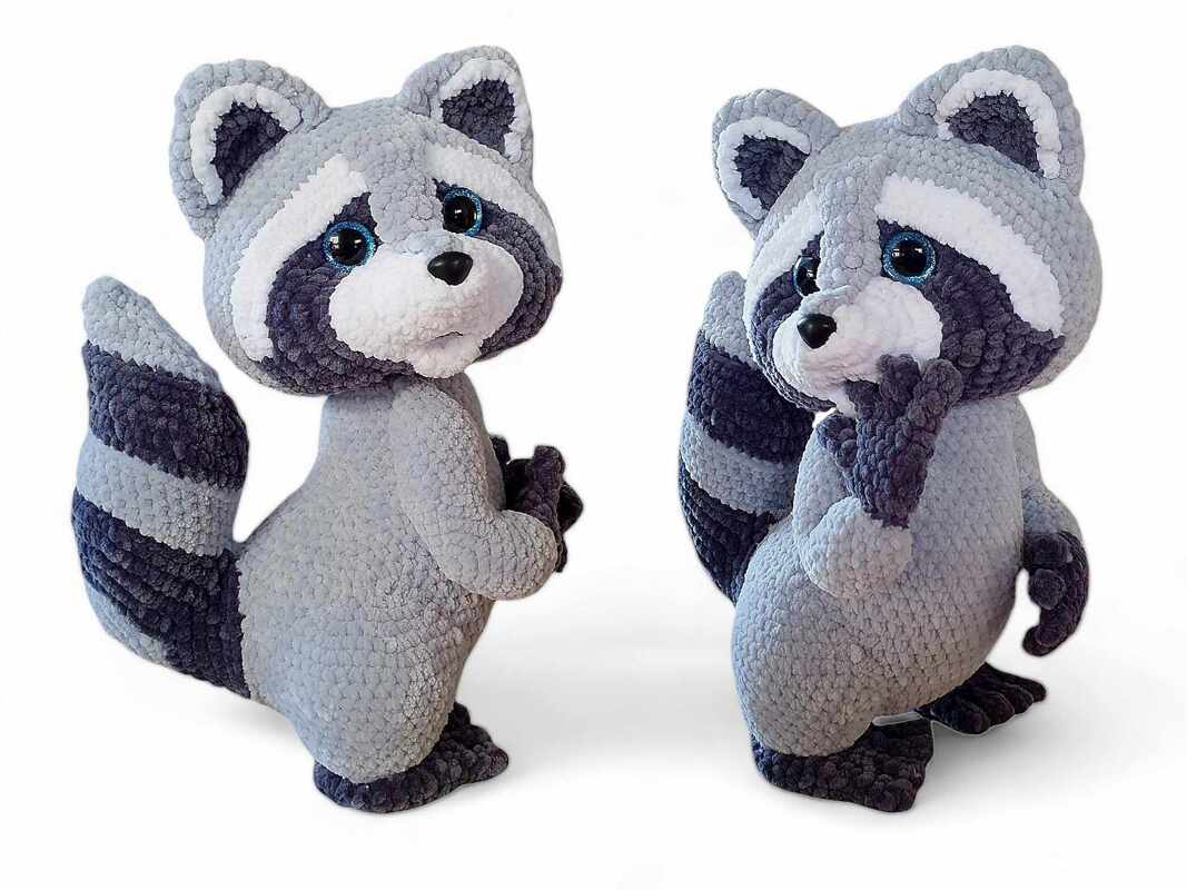 Zwei gehäkelte Amigurumi-Waschbären in Grau mit gestreiftem Schwanz und blauen Augen vor weißem Hintergrund.