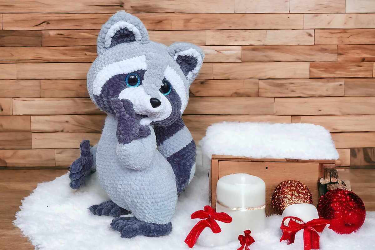 Handmade gray and white crocheted raccoon amigurumi with blue eyes on a white faux-fur rug next to candles and holiday ornaments.