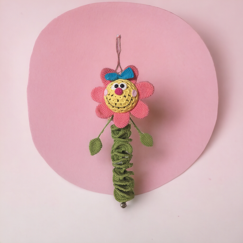 Gehäkelte hängende Blume mit rosa Blüten, gelbem Gesicht, blauem Schleifchen und grünem Spiralstiel