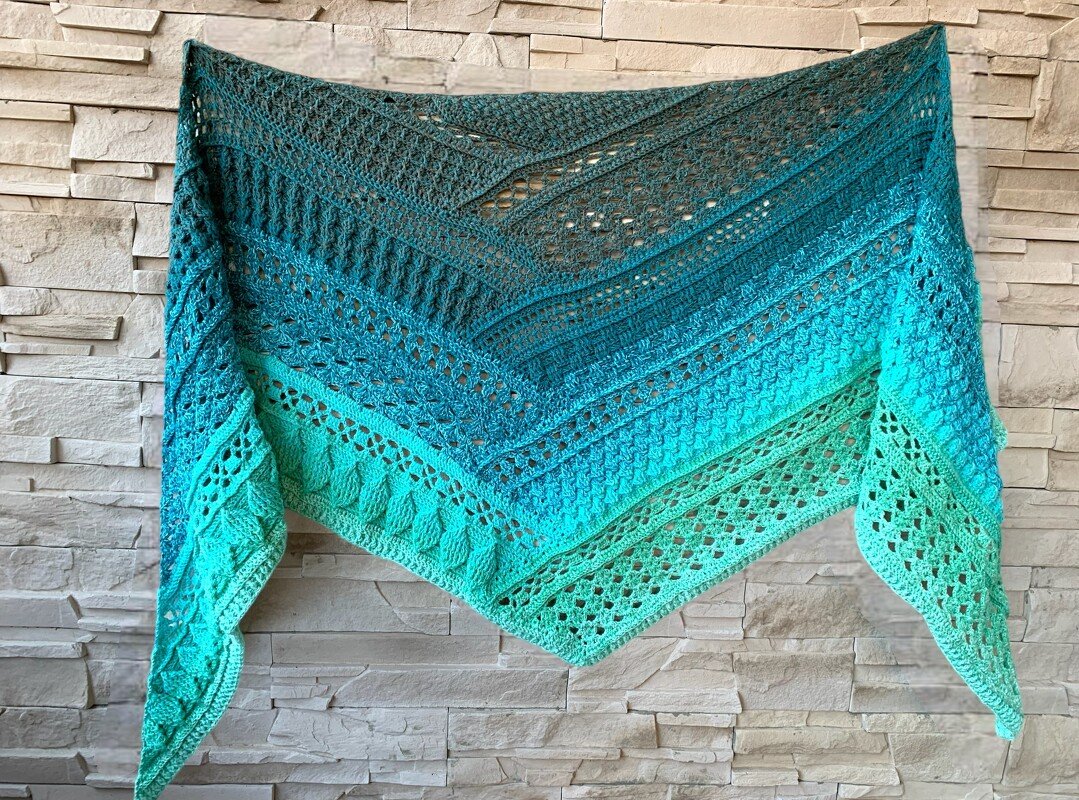 Ombre crocheted triangle shawl in a teal-to-aqua gradient, showing alternating lace and textured stitch sections, draped on a light stone wall.