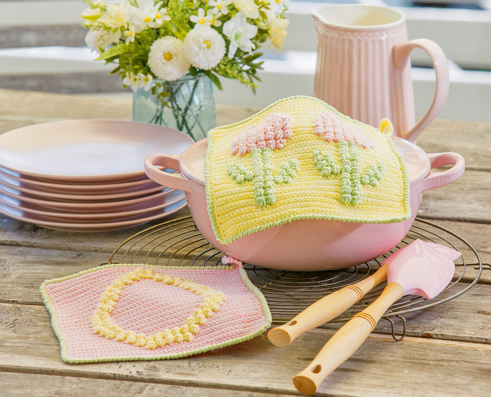 Zwei gehäkelte Topflappen in Pastell: gelb mit Kaktus-Relief auf rosa Topf, rosa Topflappen mit Kreisdetail auf Holztisch.