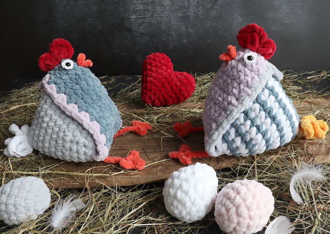Two fluffy crocheted amigurumi chickens in blue/grey and lilac/white, with a red heart and soft yarn eggs on straw.