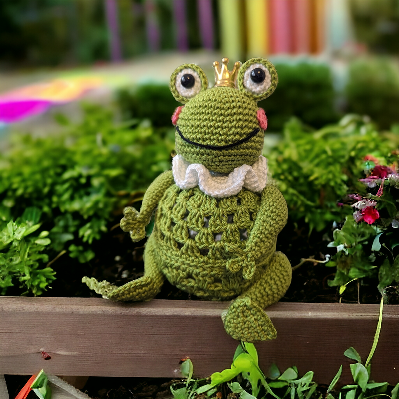 Gehäkelter grüner Frosch mit goldener Krone und weißem Rüschenkragen, sitzend auf einer Holzkante vor Moos und kleinen Blüten