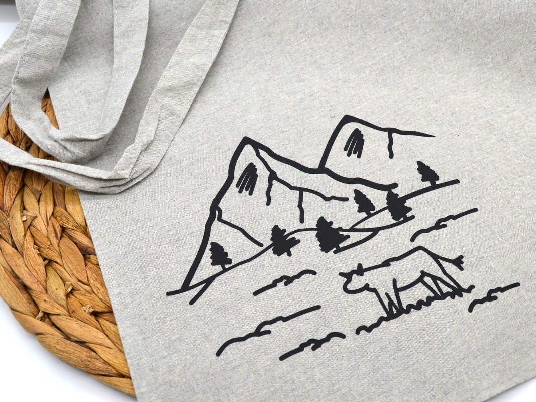 Schwarze Linienzeichnung von Bergen, Tannen und einer weidenden Kuh auf hellgrauer Stofftasche