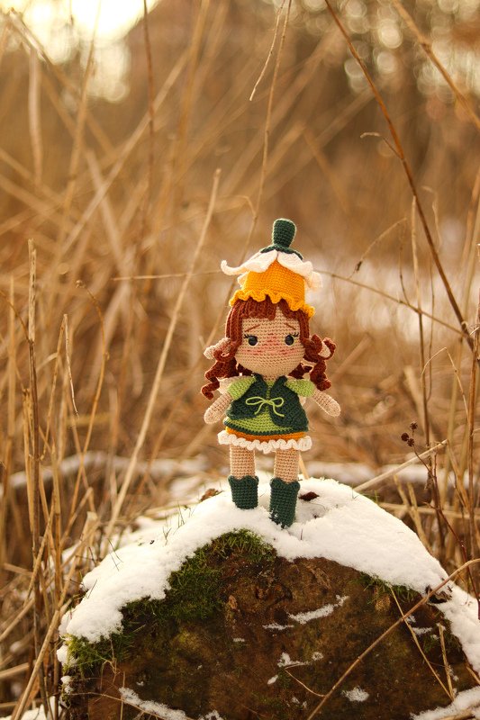 Gehäkelte Amigurumi-Blütenpuppe mit braunen Locken, grünem Kleid und gelber Blütenkappe auf einem schneebedeckten Baumstumpf im trockenen Gras.