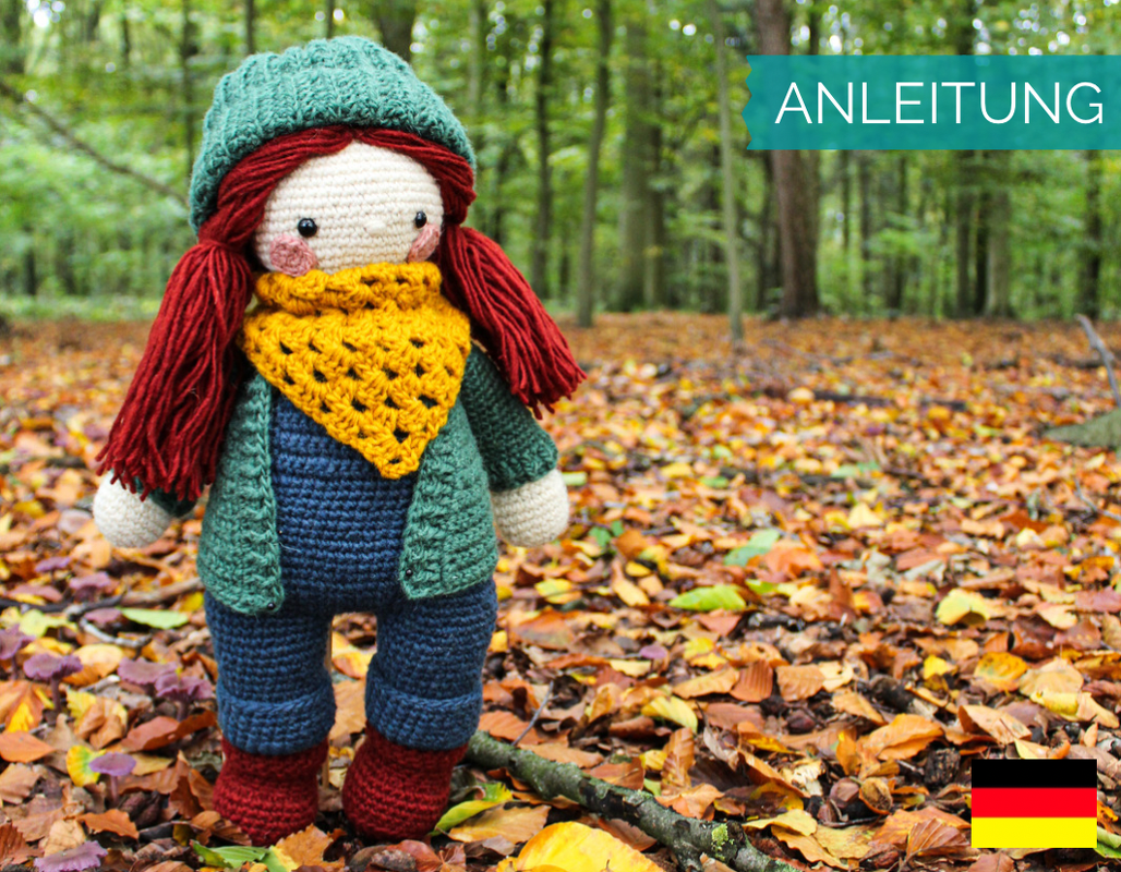 Gehäkelte Amigurumi-Puppe mit grüner Mütze, roten Zöpfen, gelbem Dreieckstuch und roten Stiefeln auf Herbstlaub