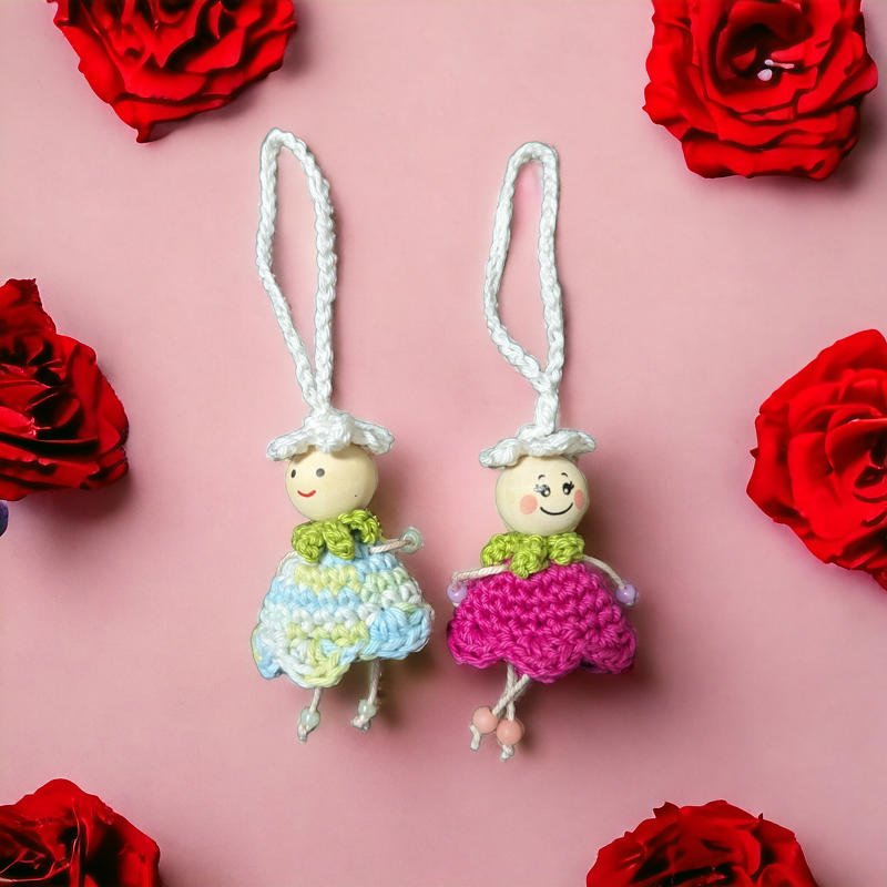 Zwei gehäkelte Püppchen‑Schlüsselanhänger mit Holzperlenköpfen und bunten Röckchen auf rosa Hintergrund, umgeben von roten Rosen