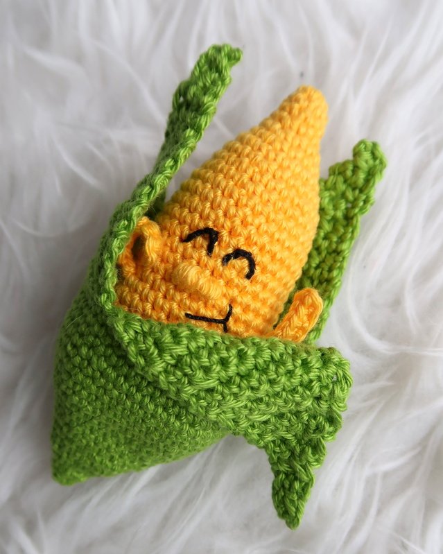 Gehäkelter gelber Maiskolben mit grüner Hülle und lächelndem Gesicht auf weißem Hintergrund.